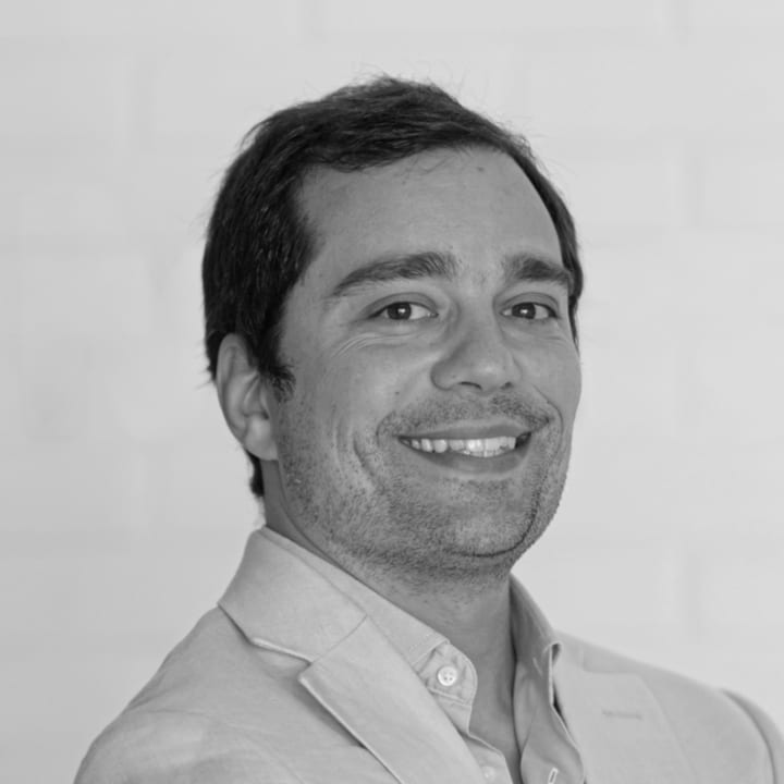 Manuel Faria, from the chest up, his body angled slightly to one side and his face turned to the camera, with an open smile. Short beard, linen jacket over an open-collar shirt. A plain, light background with nothing else in it. Black and white.